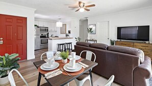 A modern living room with a brown leather sofa, a dining table set with white plates and cups, a ceiling fan, and a TV on the wall. - Walking on Sunshine – 2BR, 1BA Near Riverwalk (Fort Lauderdale)