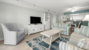 Un salon moderne avec un fauteuil gris, un téléviseur posé sur un meuble blanc, un canapé à rayures et, au fond, un coin cuisine.