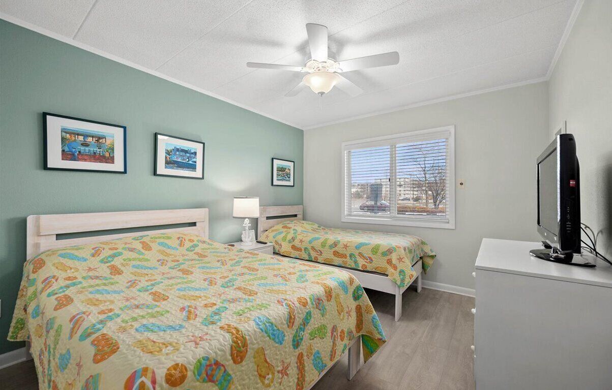 A bedroom with two beds, a ceiling fan, a TV, and framed pictures on the wall.