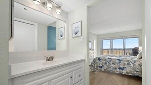 Une chambre avec un grand lit, une salle de bain avec un meuble-lavabo blanc et une vue sur la plage par la fenêtre.
