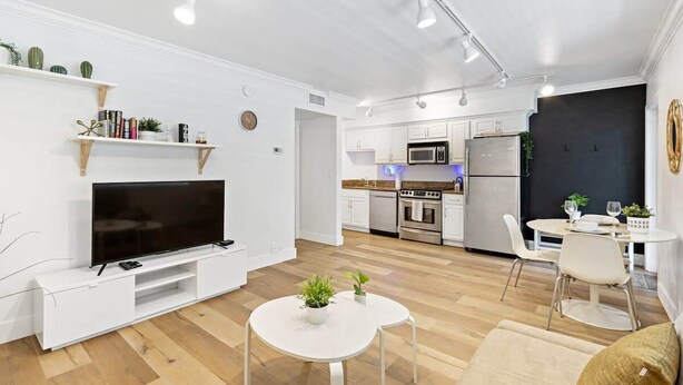 A modern living room with a flat-screen TV, white furniture, and a kitchen in the background. - Charming Waterfront Apartment – Las Olas Blvd Isle (Fort Lauderdale)