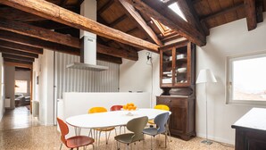 Un coin repas avec une table blanche, des chaises colorées, un plafond en bois et une vue par la fenêtre.
