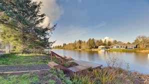 A lakeside property with a dock, a wooden deck, and a row of houses in the background. - Canal-front Home in Ocean Shores w/ Dock & Views! (Ocean Shores)