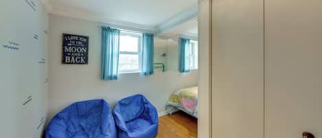 Quarto com dois pufes, uma cama com colcha colorida, uma janela com cortinas e uma parede com um aviso.