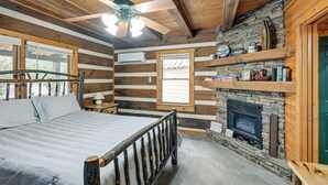 Une chambre située dans une cabane en rondins, avec un lit, une cheminée en pierre, un ventilateur de plafond et des étagères en bois.