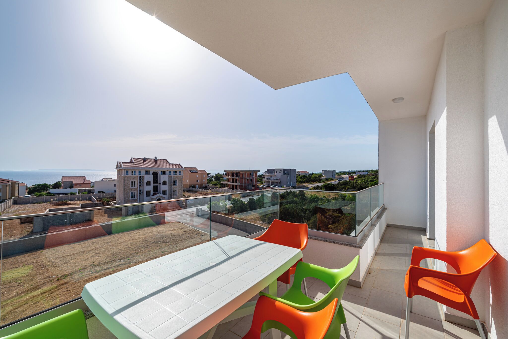 En balkon med bord og stole, fri udsigt til havet og bygninger i baggrunden.