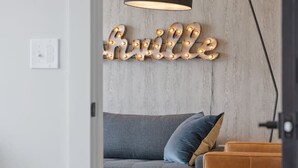 A modern living room with a grey sofa, a brown side table, and a wall-mounted light fixture spelling out "hville." - 2br condo near Vanderbilt with pool and gym (Nashville)