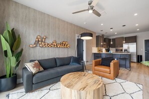 A modern living room with a grey sofa, a wooden coffee table, and a leather armchair. There is a kitchen area with dark cabinets and a central island. A large plant is visible in the corner.