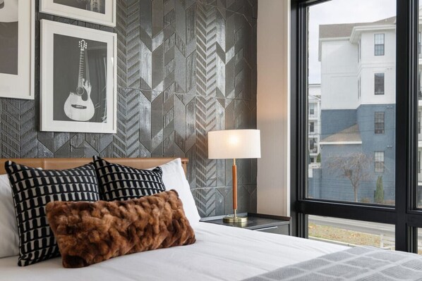 A hotel room with a bed, a nightstand with a lamp, and a window with a view of buildings.