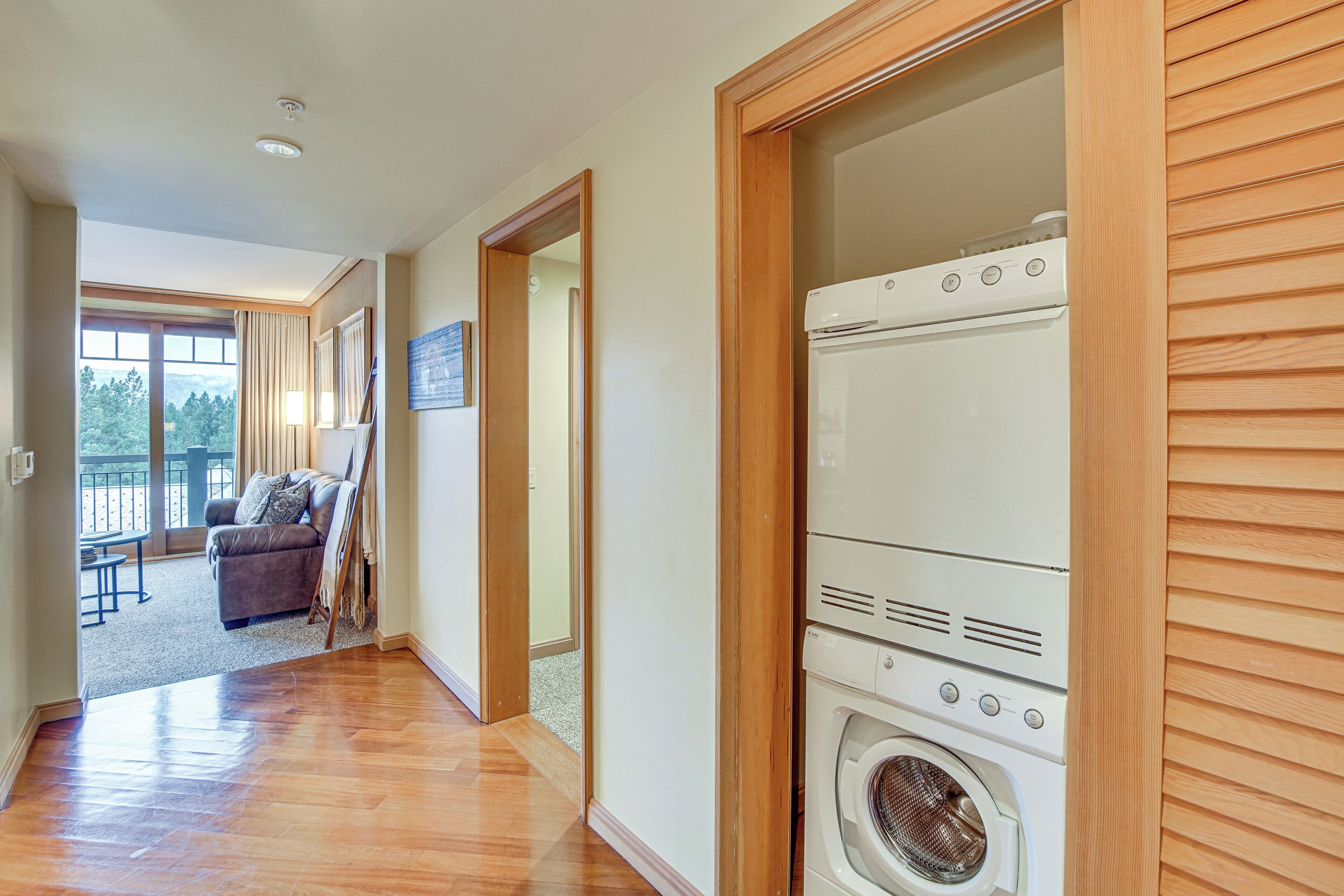 A laundry room with a washer and dryer, wooden walls, and a view to a balcony with a sofa and a painting.