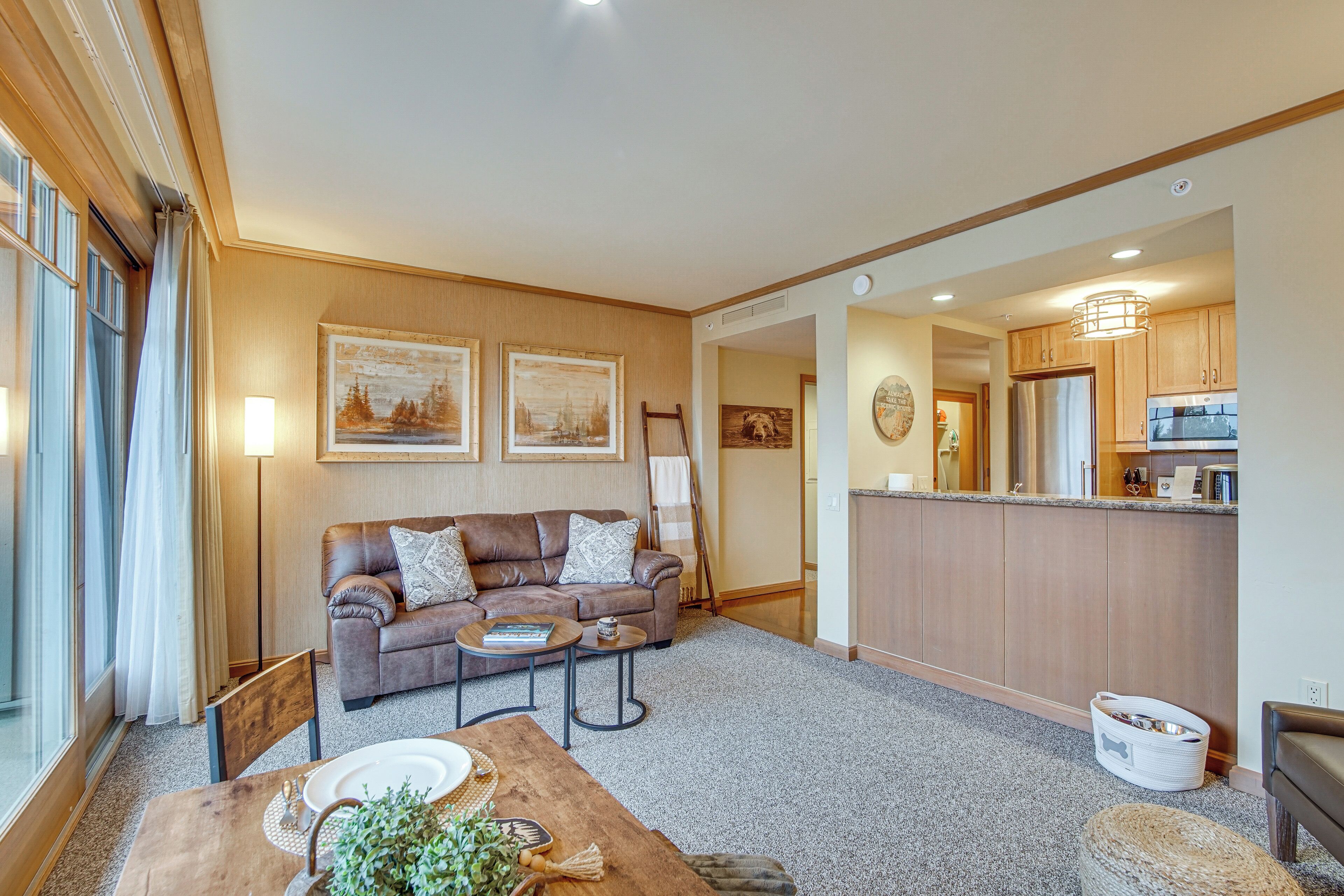 A living room with a sofa, coffee table, and dining table. There is a kitchen area with wooden cabinets and a refrigerator.