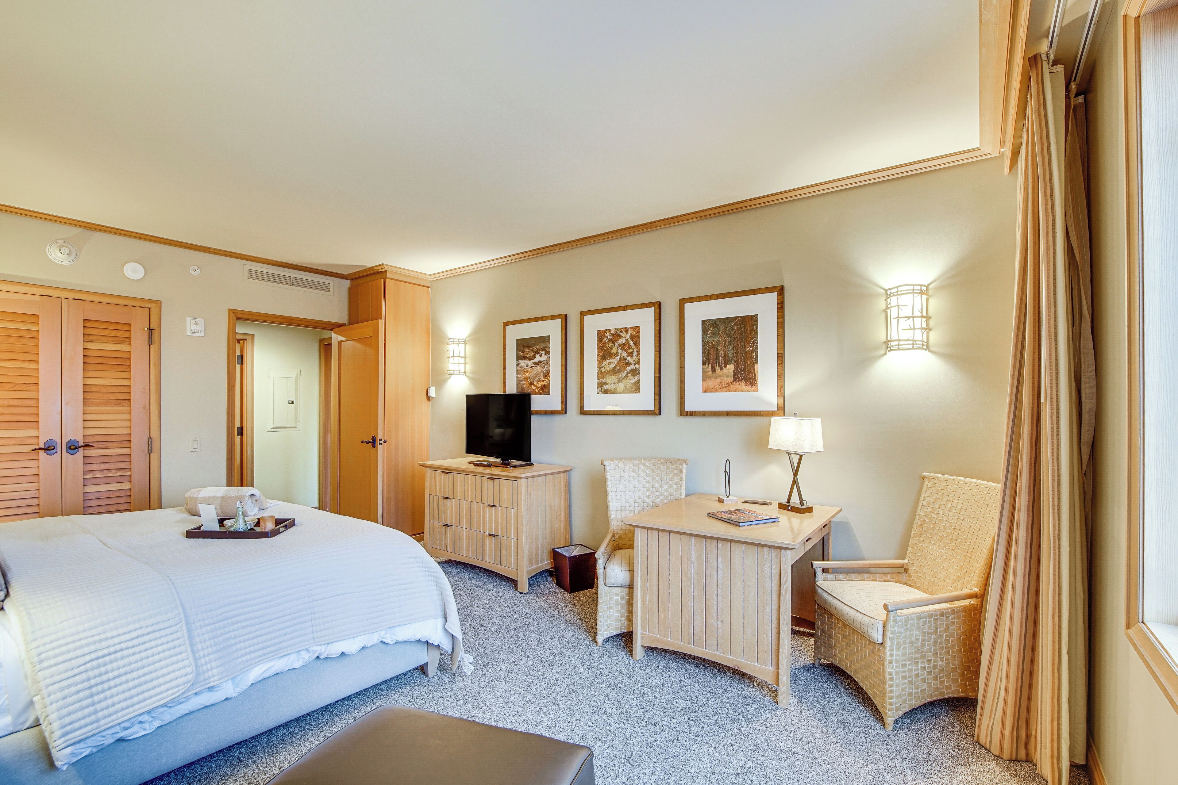 A hotel room with a large bed, a desk with a lamp, a chair, a TV on a wooden cabinet, and framed pictures on the wall.