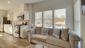 Una cocina moderna con armarios blancos, microondas integrado y estufa a gas. Hay un sofá color beige con cojines estampados frente a amplias ventanas.