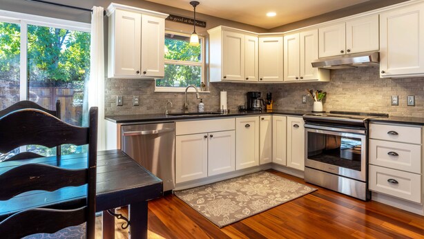 A modern kitchen with white cabinets, a stainless steel stove, and a window with a view of trees. - Devon Court 2 Bedroom Home by RedAwning (Hood River)