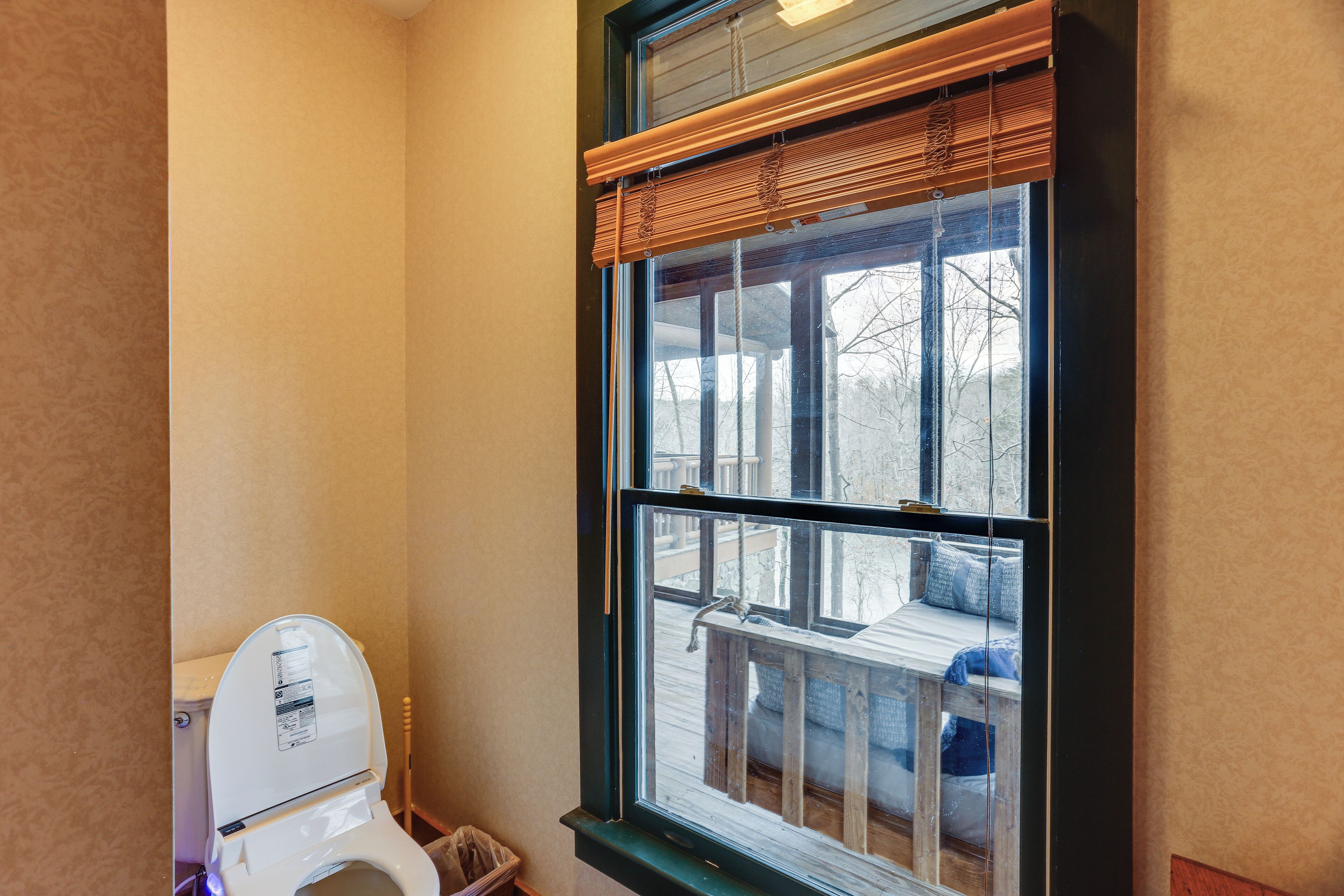 A bathroom with a toilet, a window with blinds, and a view of a deck outside.