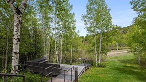 A wooden deck with metal railings, surrounded by birch trees and a grassy area with yellow flowers. - Contemporary Townhome With Ski-in/ski-out Access by The Woodrun V Townhouses by RedAwning (Snowmass Village)