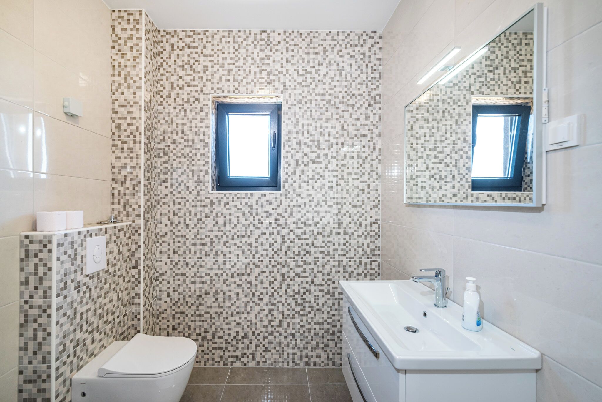 Ein Badezimmer mit Toilette, Waschbecken mit Spiegel und Duschbereich mit Mosaikfliesen.
