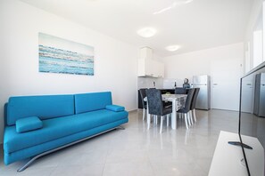 A modern living room with a blue sofa, a dining area with a table and chairs, a kitchen with white cabinets, and a large painting on the wall. - Deluxe Villa Laura 6a (Novalja)