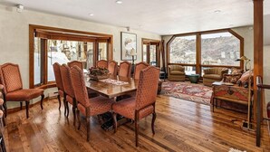 Un'ampia sala da pranzo con un tavolo in legno e sedie abbinate, grandi finestre con vista su un paesaggio innevato e un'area salotto accogliente con un tappeto decorato e cuscini.