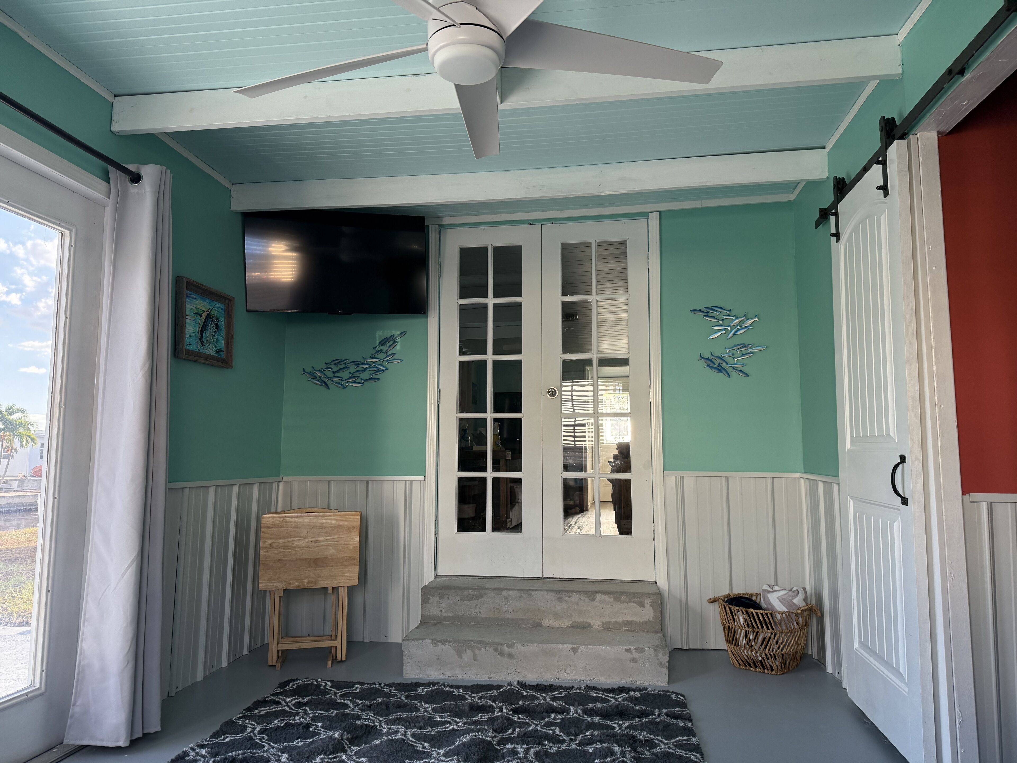 A room with teal walls, a ceiling fan, a wooden easel, a black patterned rug, and a door leading to another room.