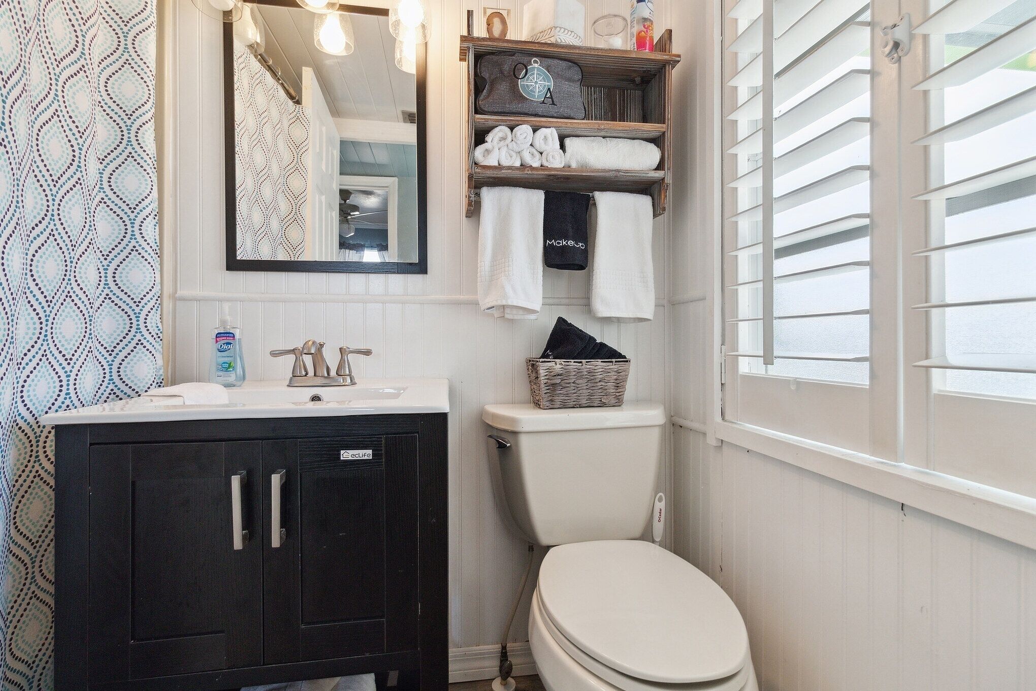 Une salle de bain avec des toilettes blanches, un meuble noir, un miroir et une étagère contenant des serviettes et des articles de toilette.