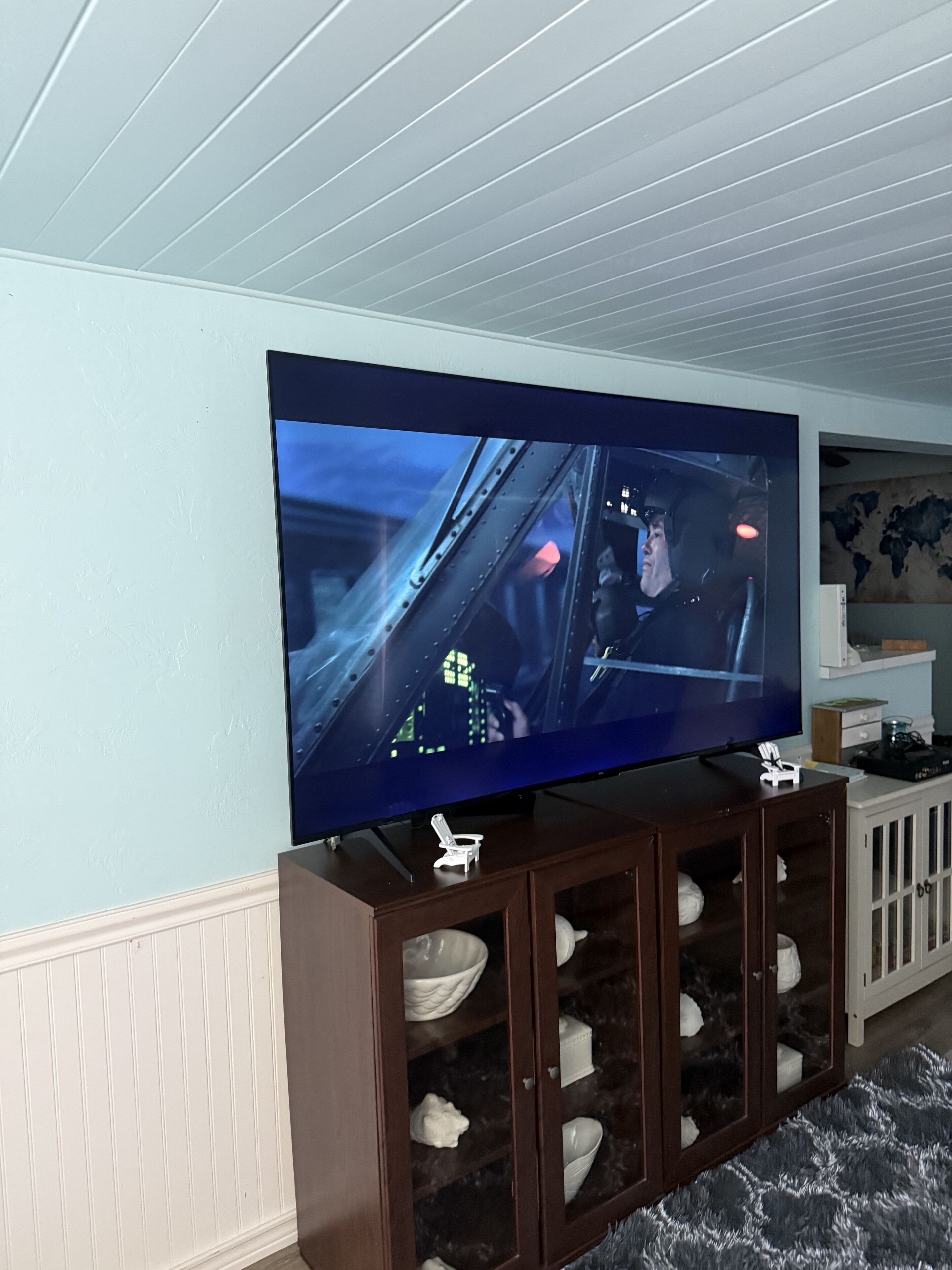 A room with a large flat-screen TV mounted on the wall, a wooden cabinet with shoes, and a patterned rug.