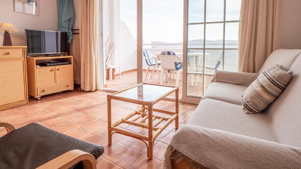A cozy living room with a sofa, a coffee table, and a TV on a wooden cabinet. There is a balcony with chairs and a view of the sea. - Casa Noray         . (La Isleta del Moro)