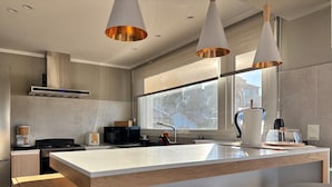 A modern kitchen with a large island, pendant lights, and a view of the outdoors through a large window. - Casa Cocó (San Carlos de Bariloche)