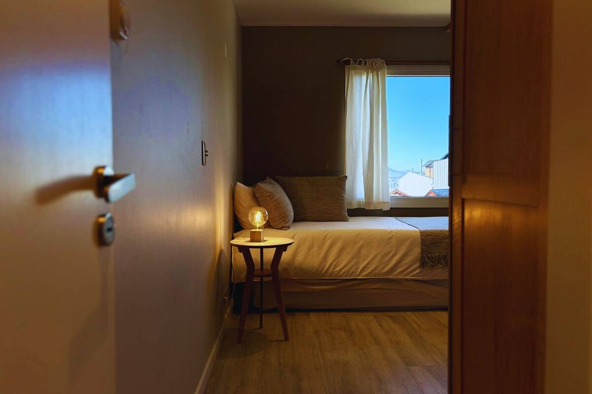 Un dormitorio con cama, una mesita de noche con lámpara, una ventana con cortinas y vistas a edificios.