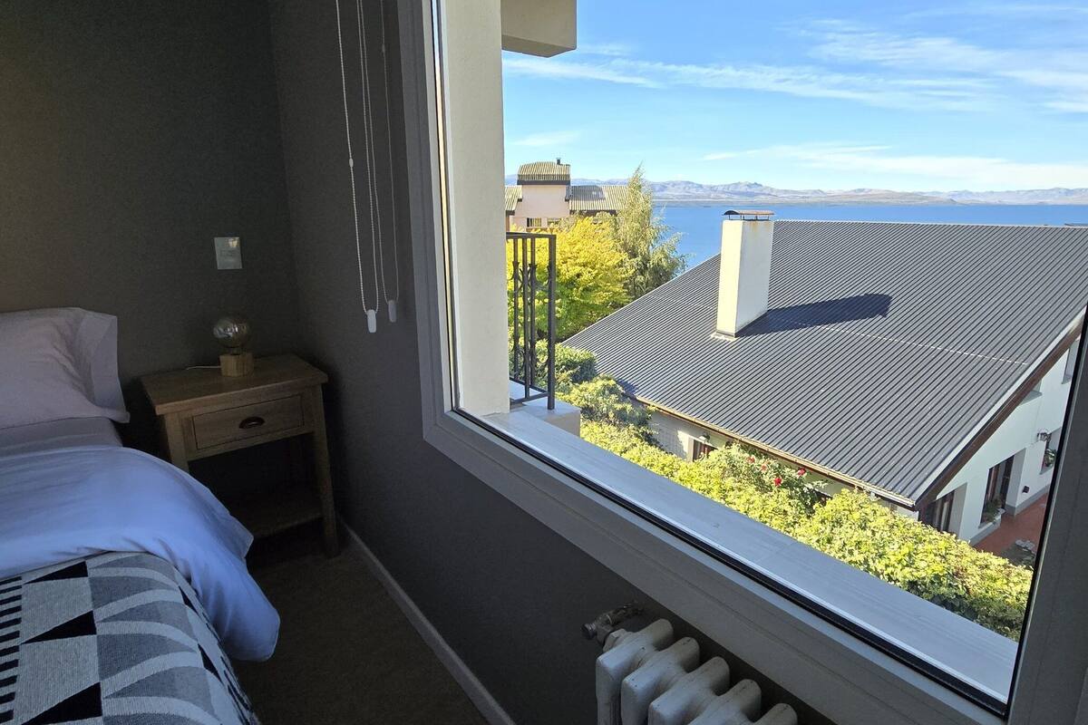 Un dormitorio con un ventanal grande que da a una zona residencial y un cuerpo de agua.