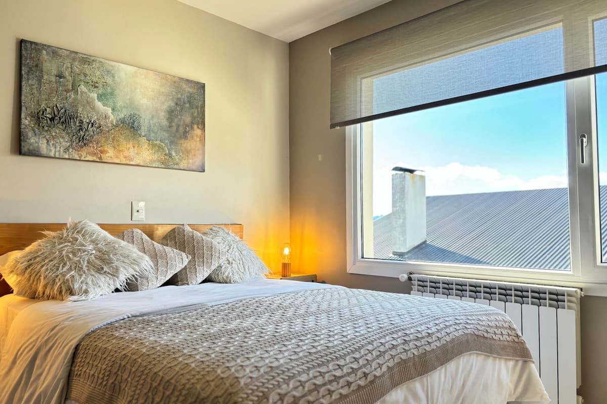 Un dormitorio con una cama grande, un cuadro en la pared y una ventana con vista a edificios.