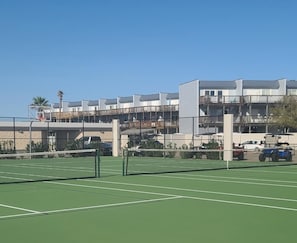 Cancha de tenis con una red en primer plano, rodeada por un edificio con varios balcones y un estacionamiento al fondo.