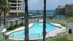 A pool area with palm trees, a fenced pool, and a view of the ocean and buildings in the background. - Lounge around our Texas size pool, barbecuing, and play tennis.  Enjoy the sunse (Port Aransas)