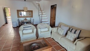 Amplia sala de estar con piso de baldosas, una mesa de centro de madera, dos sillones blancos, un sofá beige con cojines estampados, una mesa de comedor con sillas y una escalera que lleva a un piso superior.