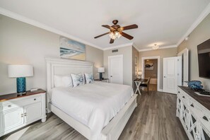 Un dormitorio con una cama grande, ventilador de techo, un cuadro del océano y una zona de comedor con mesa y sillas.