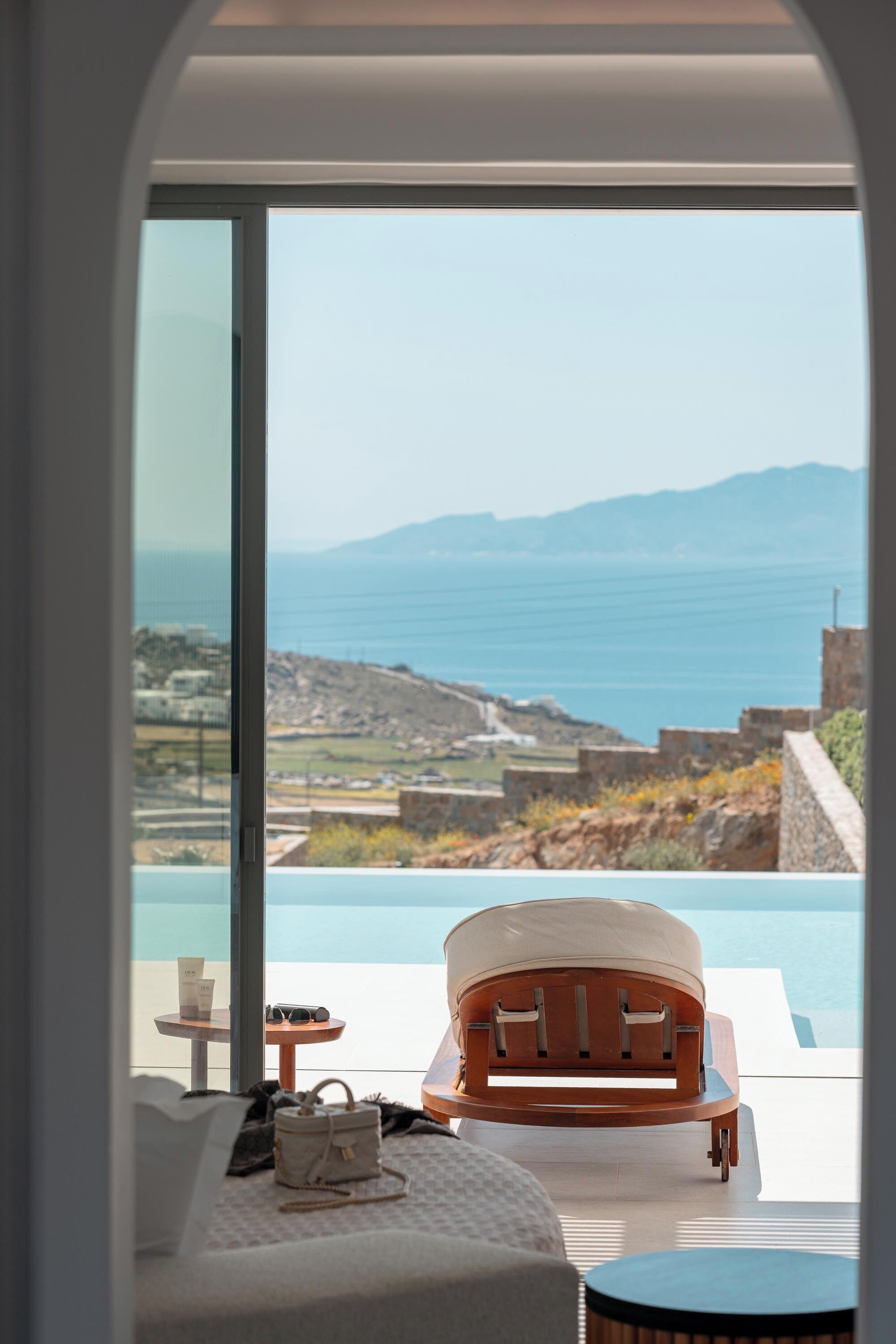A modern room with a large window overlooking a coastal landscape, a lounge chair, and a small table with a cup and sunglasses.