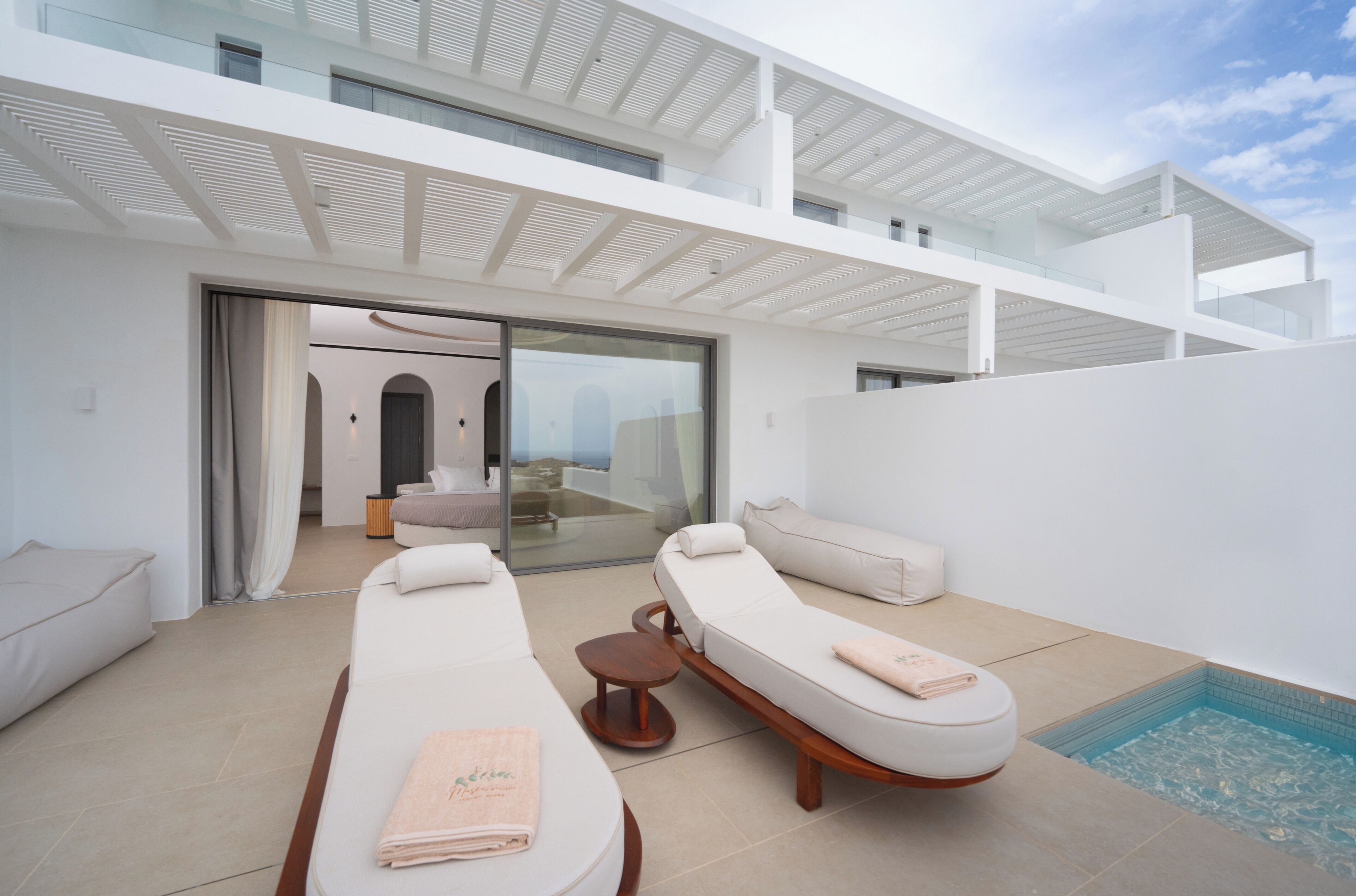 Ein moderner Außenbereich mit Liegefläche, zwei Sonnenliegen und einem kleinen Tisch, eine Glastür zu einem Innenraum und ein Pool, der durch das Glas sichtbar ist.