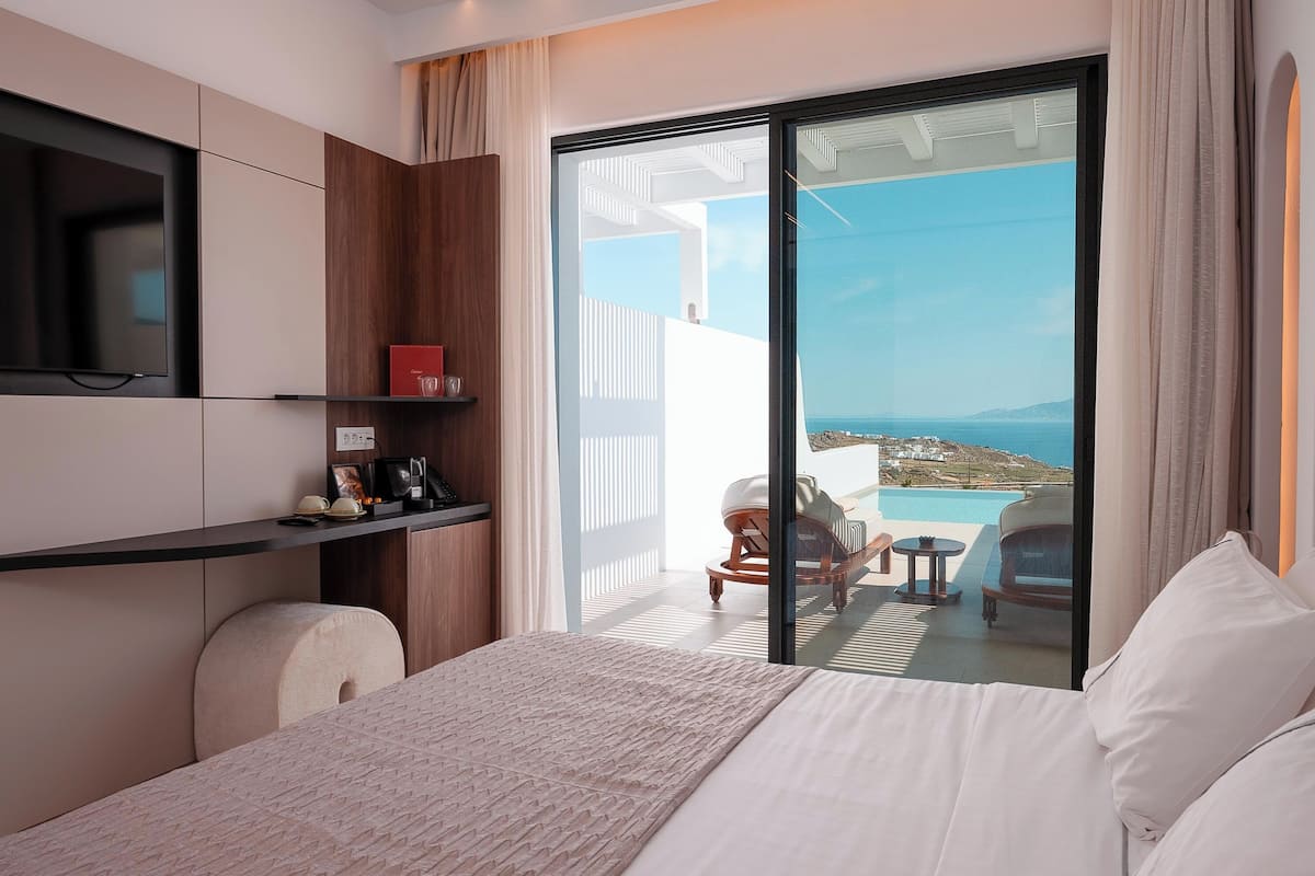 A hotel room with a bed, a TV, a small shelf with items, and a balcony with a pool and chairs.