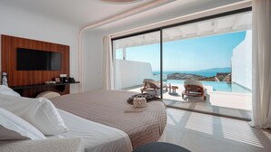 A modern hotel room with a large bed, a flat-screen TV mounted on a wooden panel, a desk, and a view of a pool and ocean. - Muse of Mykonos  (Mykonos)