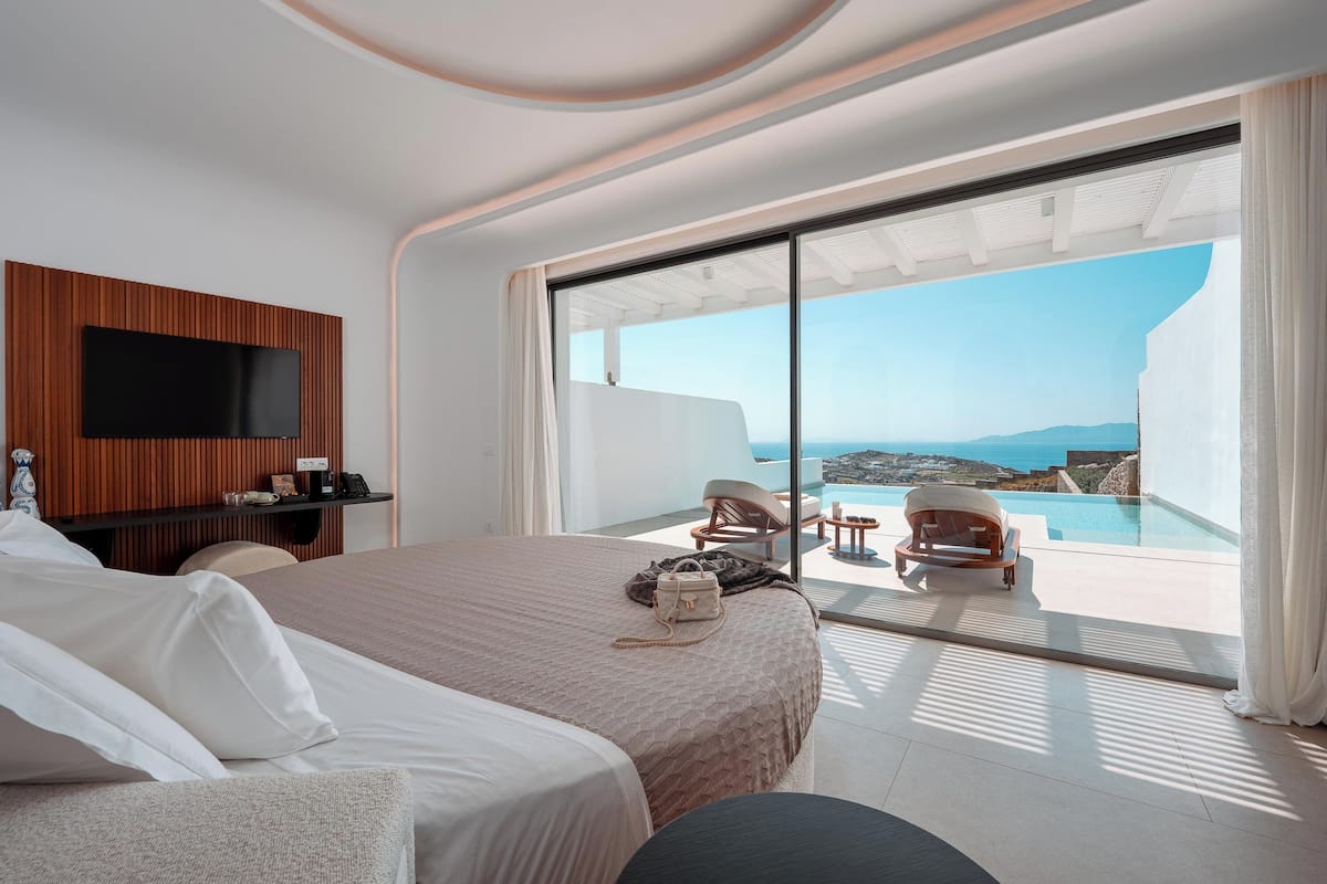 A modern hotel room with a large bed, a flat-screen TV mounted on a wooden panel, a desk, and a view of a pool and ocean.