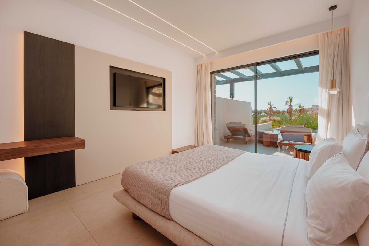 A modern hotel room with a large bed, a flat-screen TV mounted on the wall, a seating area with a table and chairs, and a balcony with a view of the landscape.