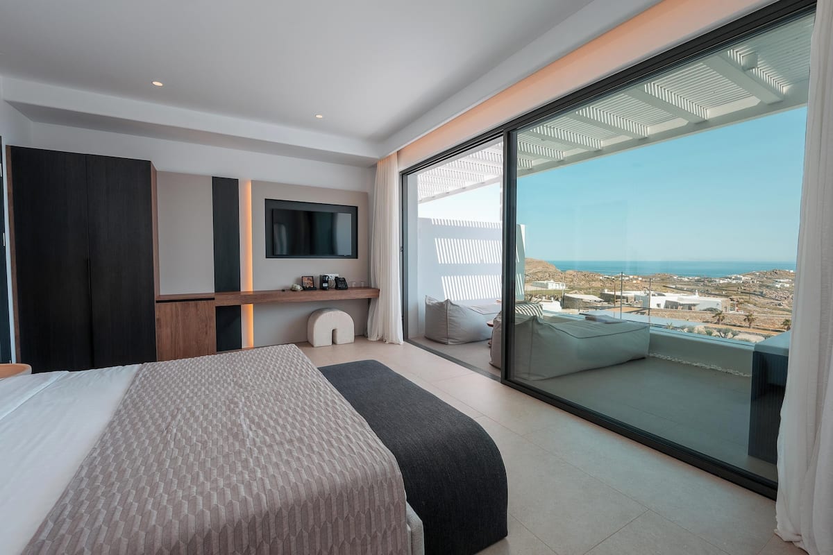 A modern hotel room with a large bed, a desk, a TV, and a balcony with a city view.