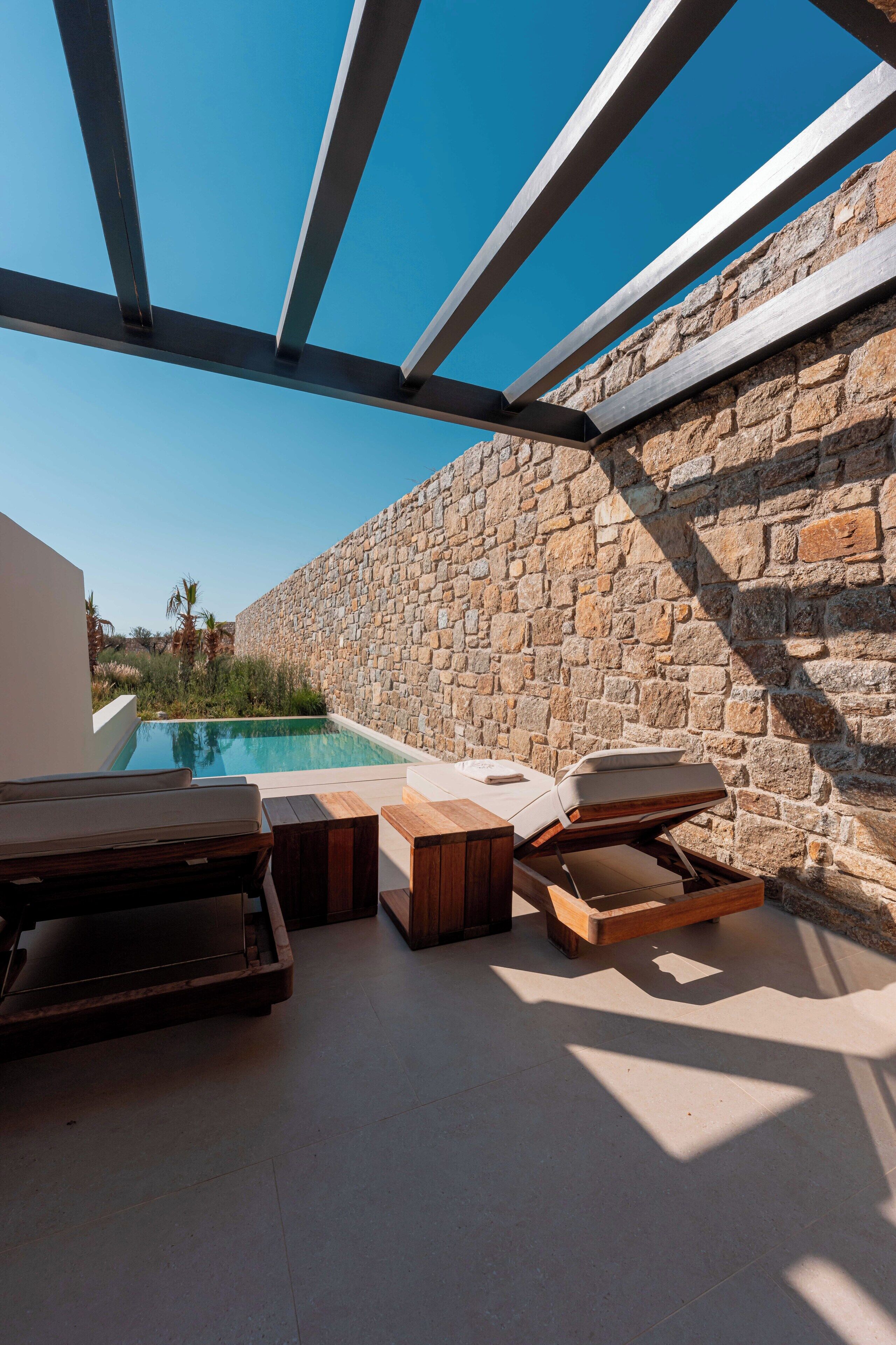 An outdoor pool area with stone walls, wooden lounge chairs, and a pergola structure.