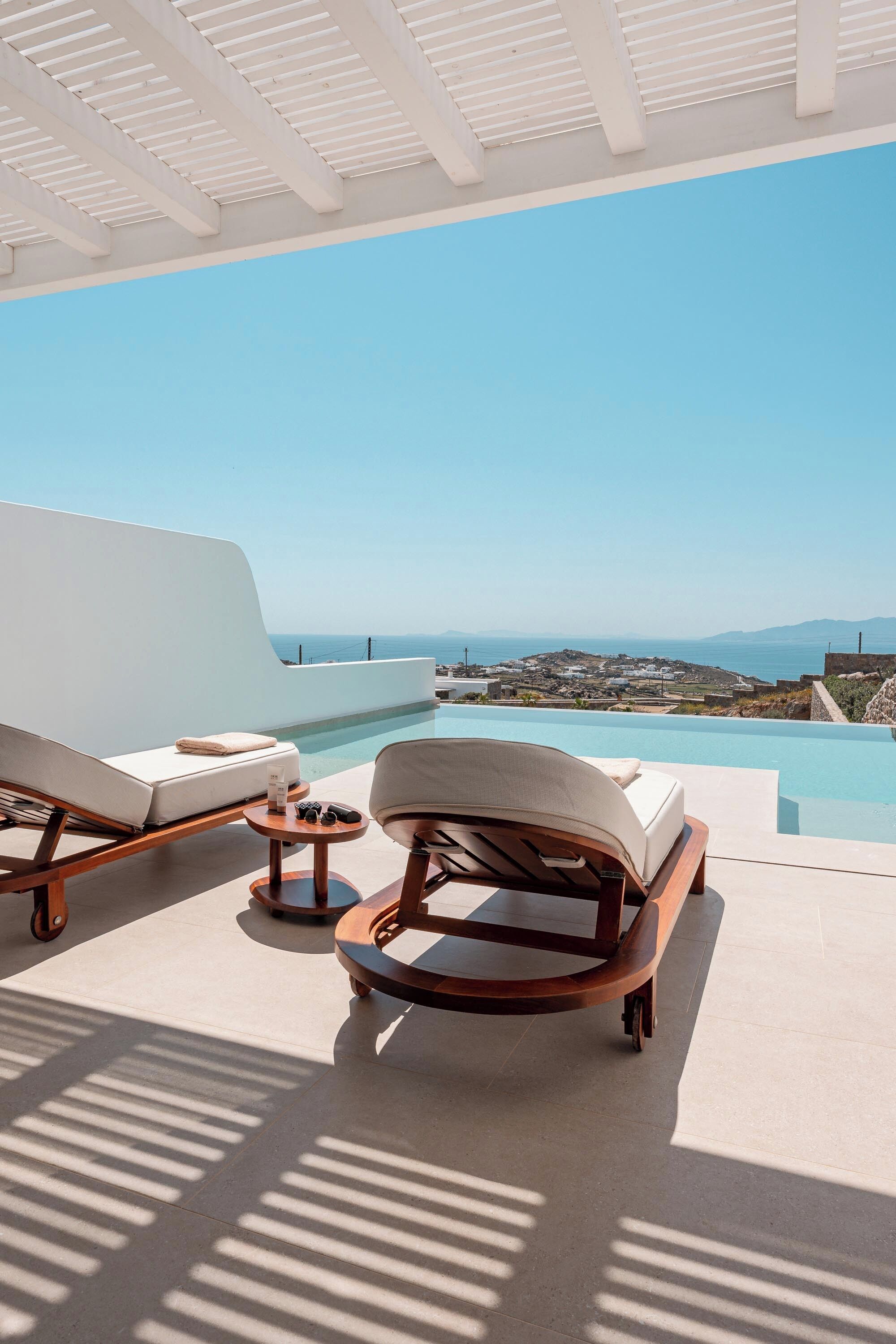 A sunlit outdoor lounge area with two white cushioned chairs and a small round table, overlooking a clear blue sky and a pool.