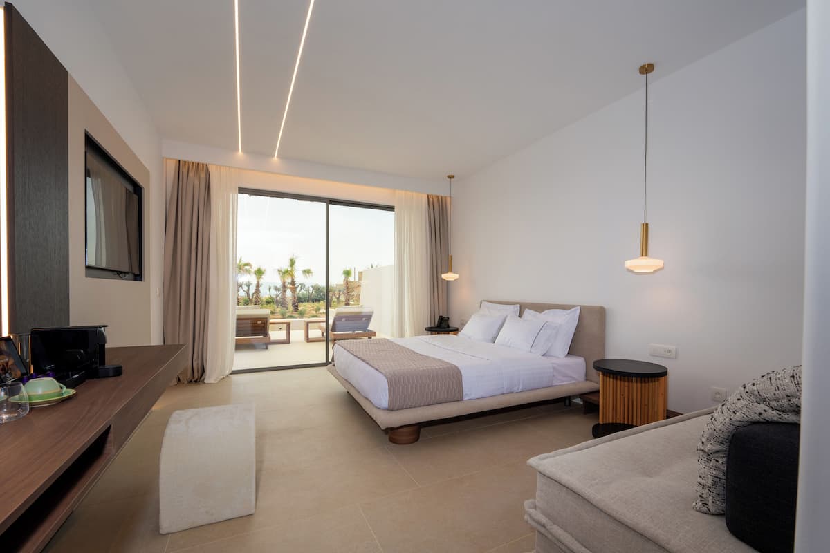 A modern hotel room with a bed, a sofa, a desk, and a view of the outdoors.