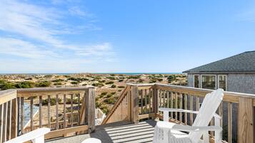 A wooden deck with two white Adirondack chairs, a small table, and a view of the beach and ocean.