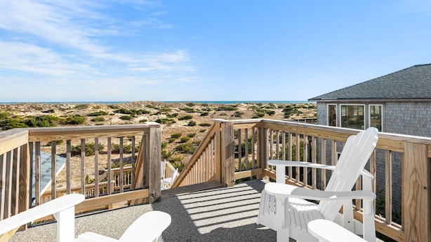 A wooden deck with two white Adirondack chairs, a small table, and a view of the beach and ocean.