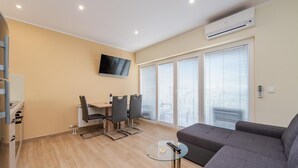 A modern living room with a grey sofa, a glass coffee table, a TV mounted on the wall, and a dining area with a table and chairs.