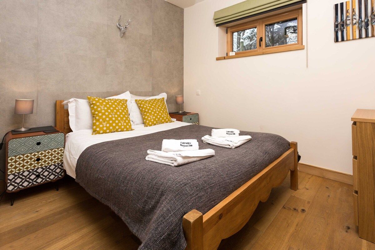 A bedroom with a wooden bed, a patterned nightstand, a window with a view, and a wall-mounted artwork.