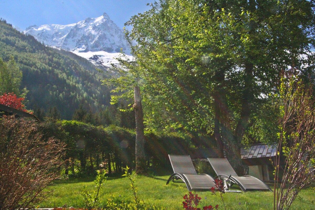 A garden with two lounge chairs, a small building, and a mountain range in the background.